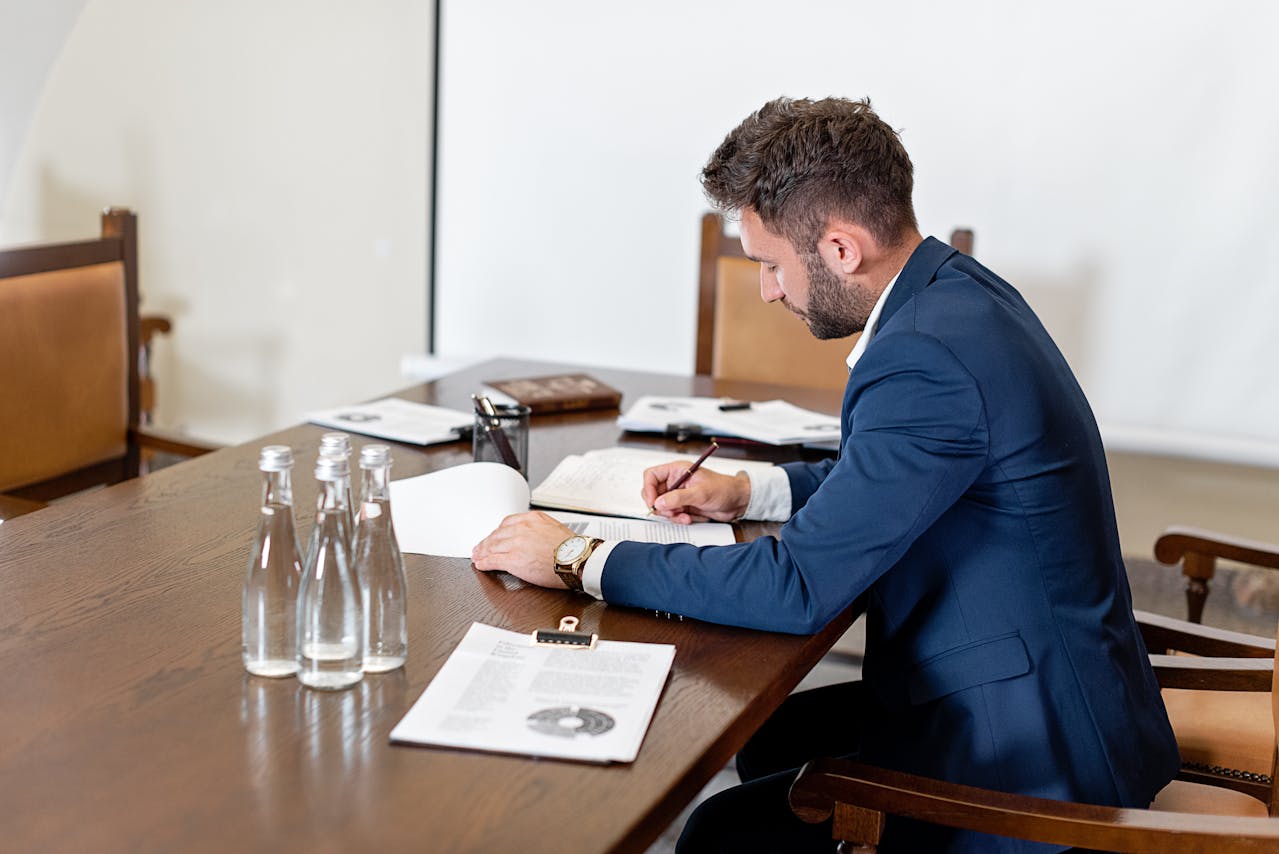 A man in a suit is writing notes at a conference table with documents and water bottles nearby.