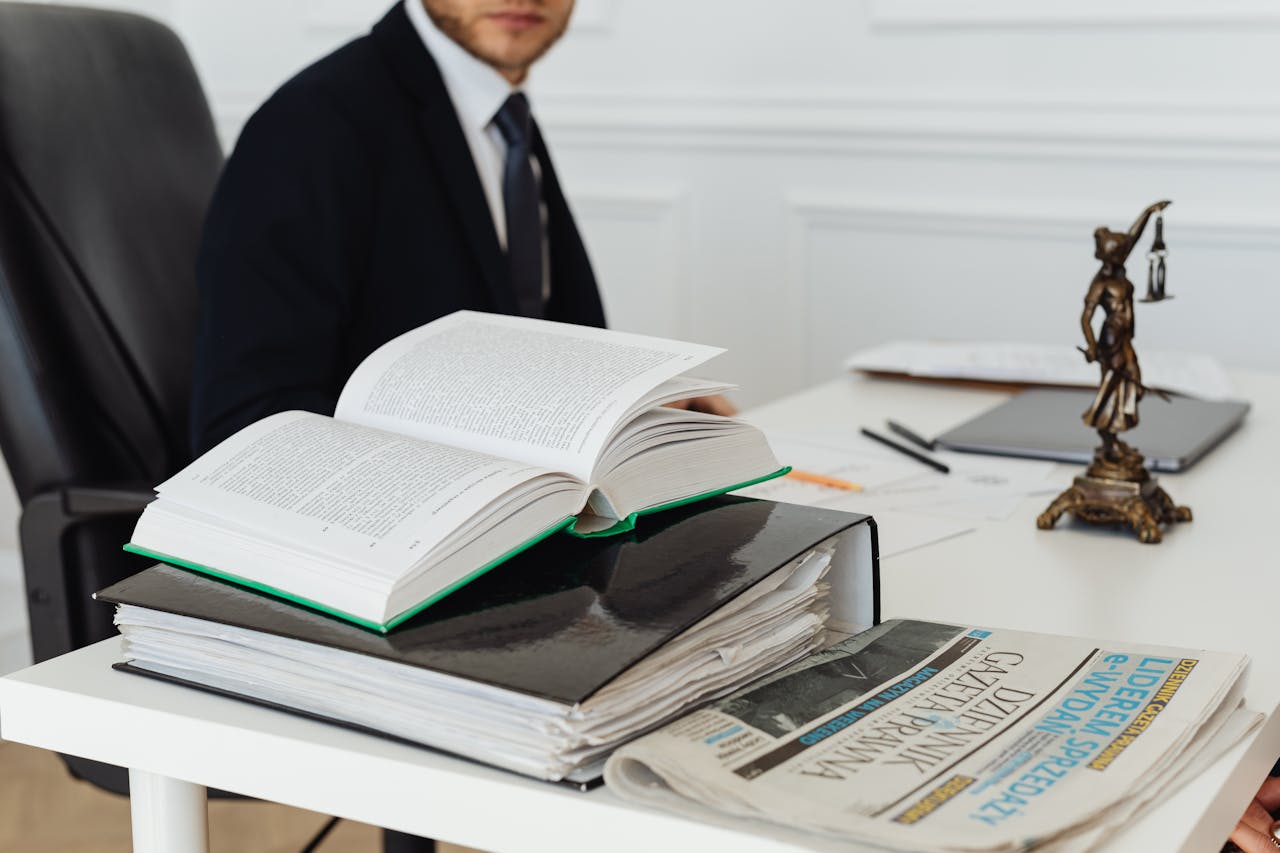 A desk with an open book, a stack of documents, a newspaper, and a statue of Lady Justice. A person in a suit is partially visible.
