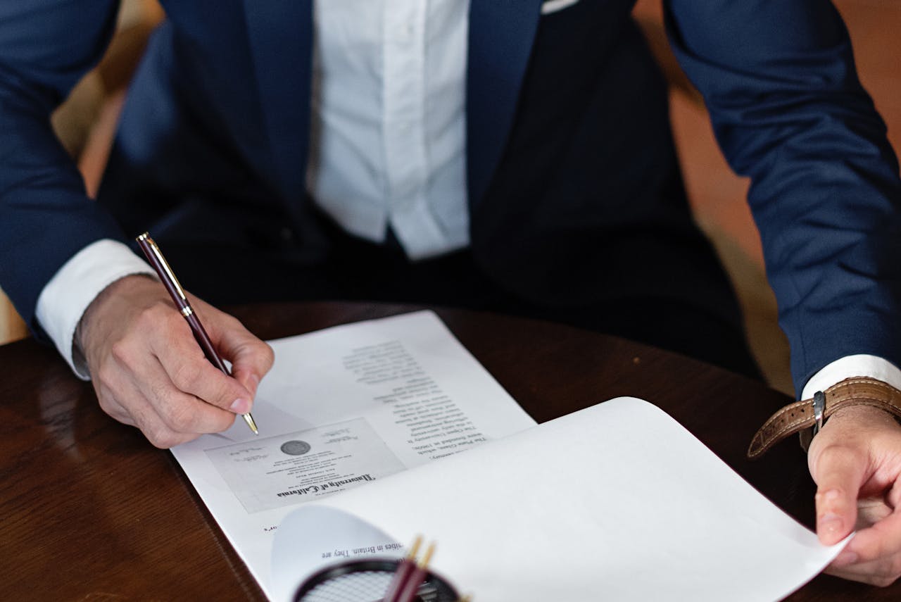A person in a suit is signing a document on a wooden table, with a pen in hand and additional papers nearby.