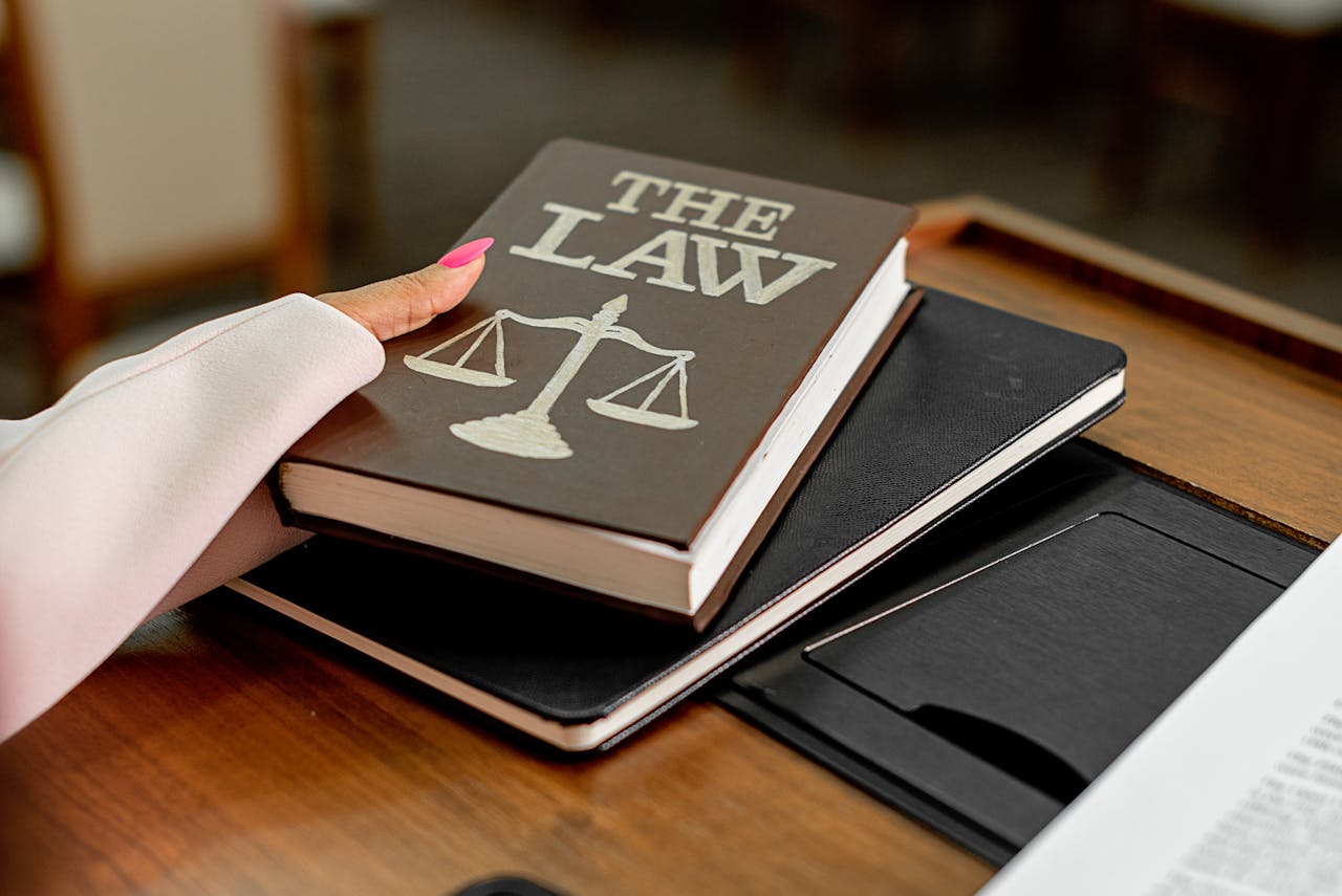 A hand with pink nails holds a book titled 'THE LAW,' resting on top of two notebooks on a wooden desk.